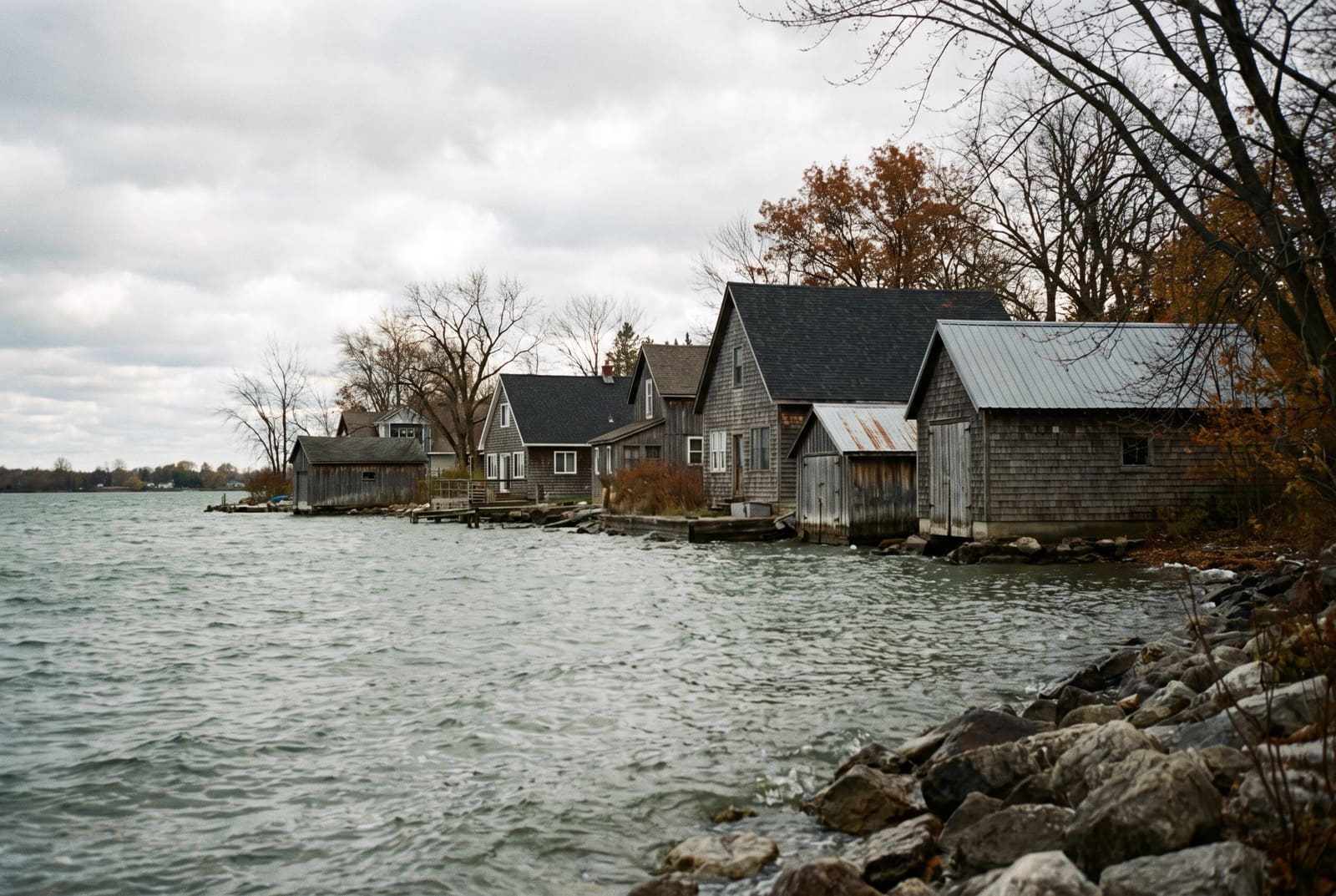Shoreline cottages and boathouses with grey shingle and metal roofs on an overcast day, rocks and choppy water in front of them.