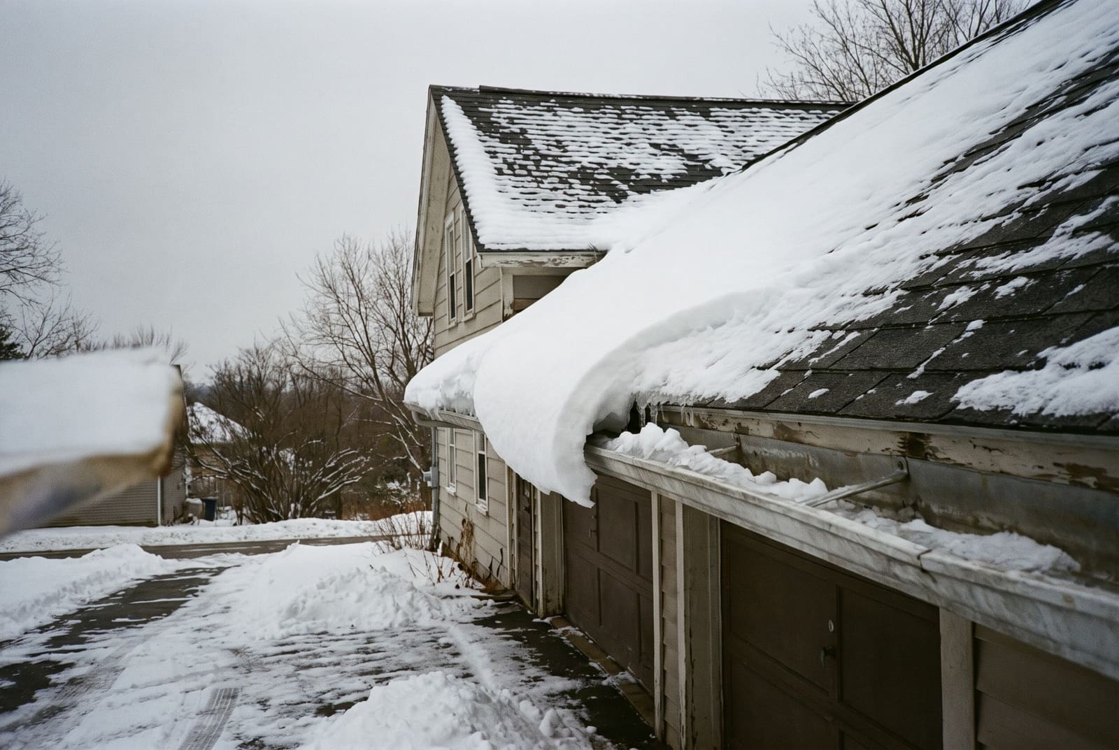 A roof eave seen from a snowy driveway, carrying a deep, rounded lip of snow that curls out over the gutter.