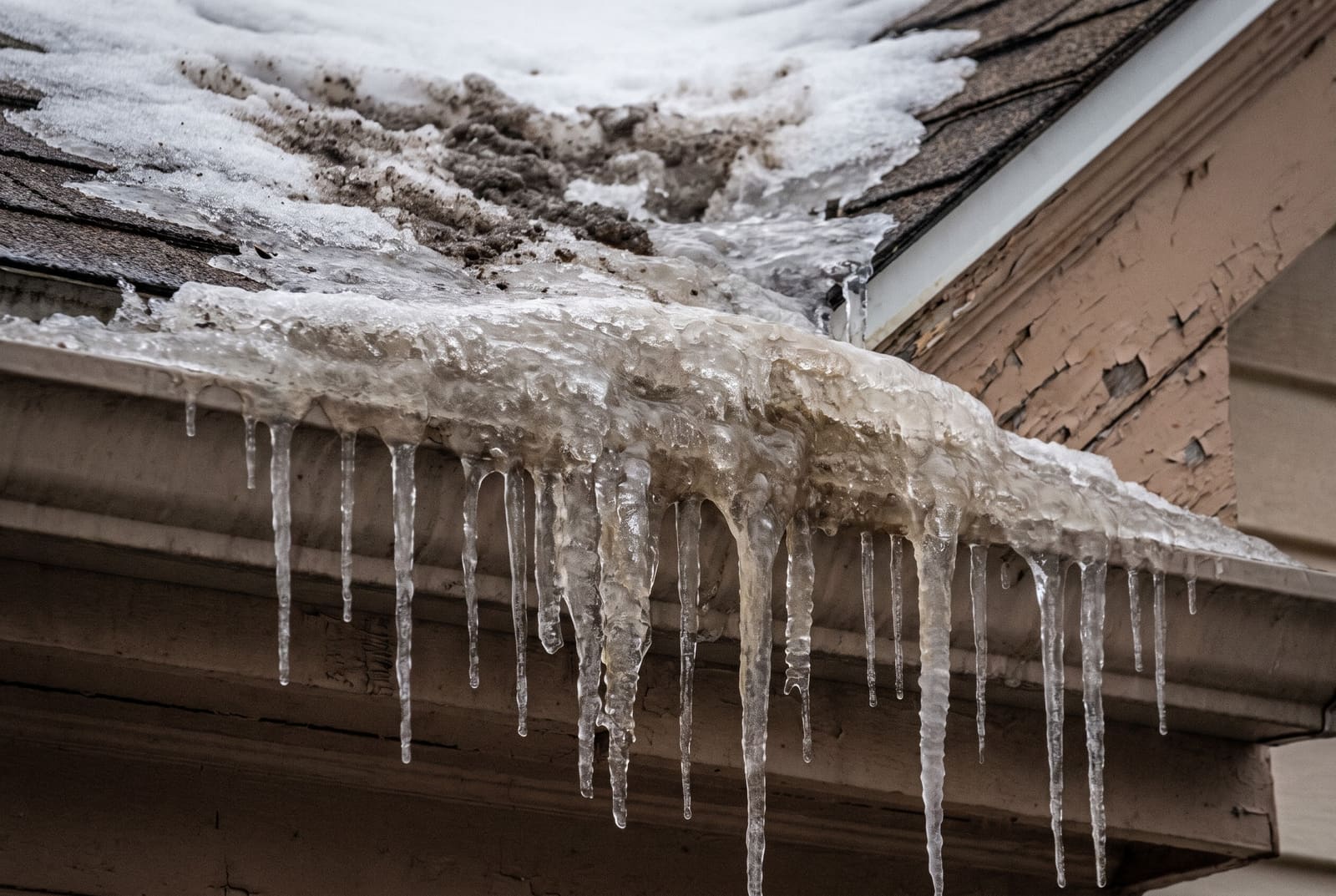 A thick ridge of ice along a roof gutter with icicles hanging from it and packed snow on the shingles above.