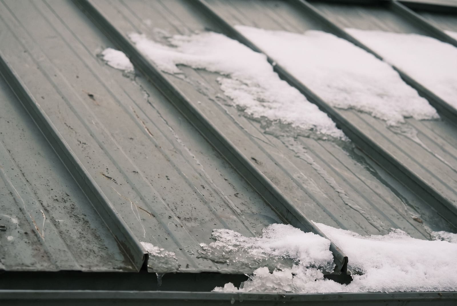 Grey-green standing-seam metal roof panels, close up, with patches of snow sliding down between the seams.