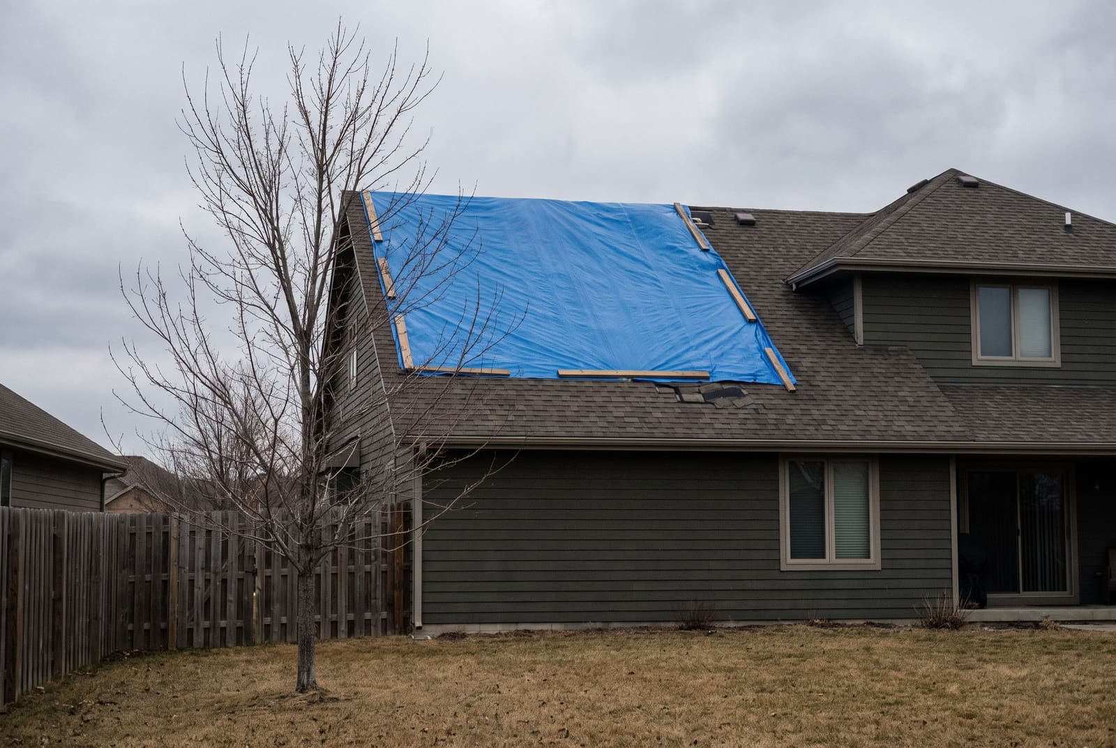 A blue tarp fastened over one slope of a shingled two-storey house, a bare tree and a wooden fence in the yard.
