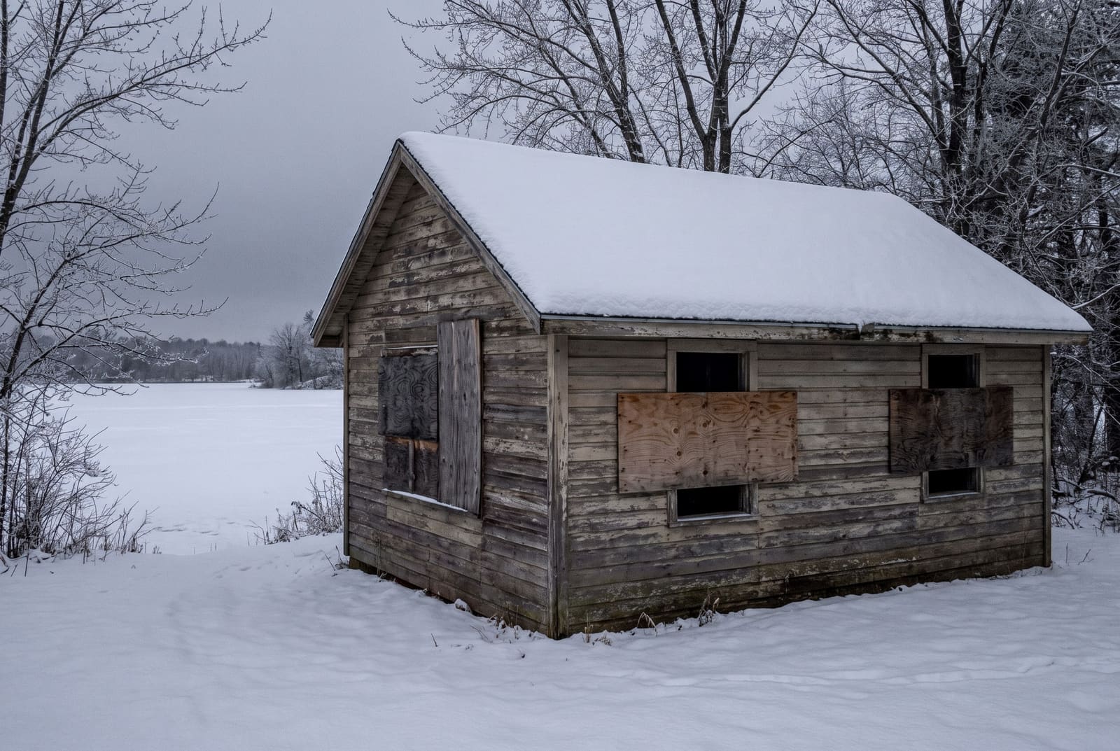 A small weathered cabin closed for the season, plywood fixed over parts of its windows, with snow on the roof and across the frozen ground.